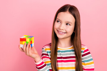 Photo of optimistic small young girl playing square toy dimensional puzzle rubik cube thinking solution isolated on pink color background
