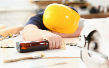 Arm of drunken worker in yellow helmet hold liquor bottle sleeping at table closeup. Manual job...