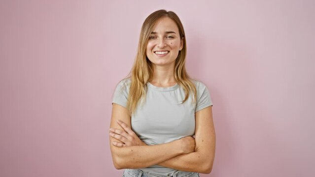 Joyful young blonde woman, cheerfully smiling, standing with crossed arms over an isolated pink background - a vibrant expression of happiness!