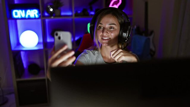 Smiling young blonde streamer woman enjoying the game, streaming live video call from her gaming room at home, engrossed in her virtual world of digital entertainment