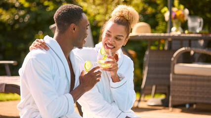 Couple Wearing Robes Outdoors Sitting Doing Cheers With Drinks By Swimming Pool On Spa Day