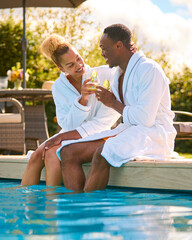 Couple Wearing Robes Outdoors Sitting Doing Cheers With Drinks By Swimming Pool On Spa Day