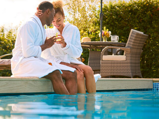 Loving Couple Wearing Robes Outdoors Sitting With Drinks With Feet In Swimming Pool On Spa Day