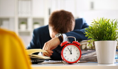 Red alarm clock shows late time closeup and tired office male clerk in suit take nap on table workplace full of exam papers. Career frustration freelance employment fail study problem low energy down