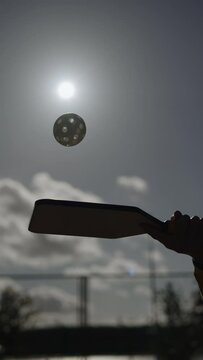 Silhouette of plastic ball with holes bouncing on pickleball racket against background of sun. Unrecognizable man hits ball while practicing pickleball in vertical video outdoor.