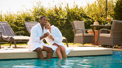 Loving Couple Wearing Robes Outdoors Sitting With Drinks With Feet In Swimming Pool On Spa Day
