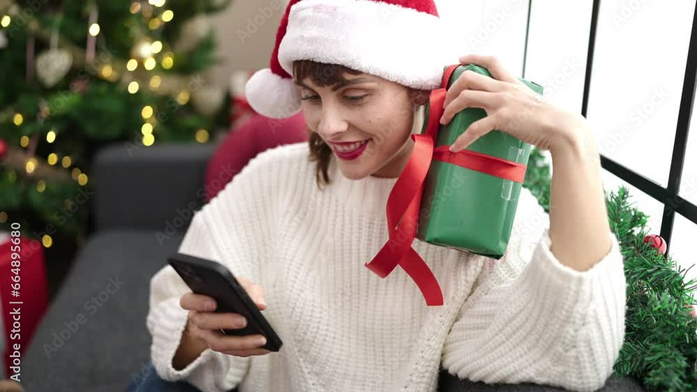 Sticker young caucasian woman using smartphone sitting by christmas tree with present at home