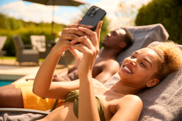 Couple On Holiday Wearing Swimming Costumes Looking At Mobile Phone On Lounger By Pool
