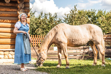 A woman in a retro blue dress with an apron is standing by a horse in the farmyard. Organic rustic...