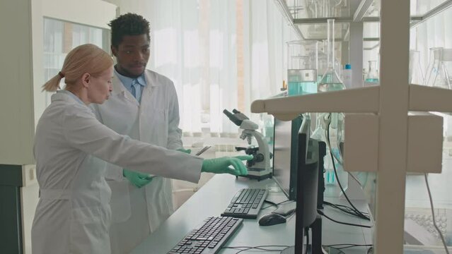 Medium shot of multiracial couple of scientists in white lab coats comparing data on computer monitors and having conversation while doing research in laboratory - Powered by Adobe