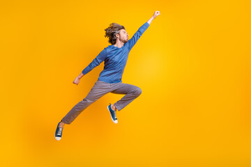 Fototapeta na wymiar Full length photo of positive cool man wear blue sweater jumping high catching arm empty space isolated yellow color background