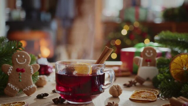 cozy composition of hot steaming mulled wine with orange slice, cinnamon stick and anise in glass mug. christmas decorations, gingerbread cookies, fir branches and flame in fireplace on background