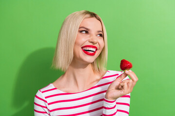 Photo of cute young girl bob blonde hair toothy hollywood smile hold red strawberry interested look novelty isolated green color background