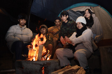 Group of Asian friends gathered around a campfire and warming their hands at night. 