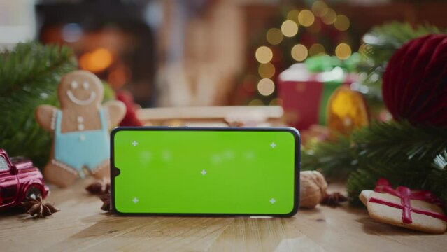 smartphone with green screen chroma key display with motion trackers on table with fir tree branches, gingerbread men and christmas decoration. burning fireplace and flashing lights on background