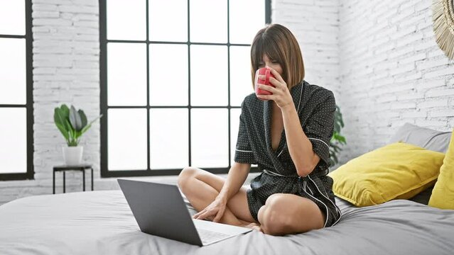 Young beautiful hispanic woman doing video call with laptop sitting on the bed drinking coffee at bedroom