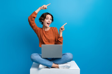 Full length photo of lovely young lady sit cube point empty space netbook wear trendy brown garment isolated on blue color background - obrazy, fototapety, plakaty