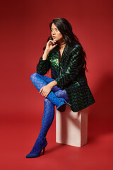 stylish asian woman in stylish green jacket with sequins and blue pantyhose sitting on red backdrop