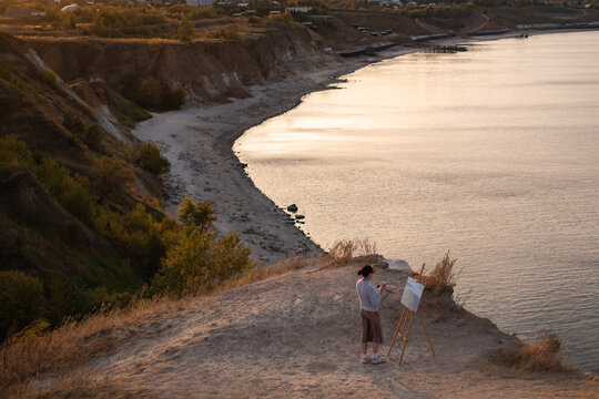 A young woman stands on a beach at sunset and paints an oil painting on an easel. The artist paints a picture of the river bank.