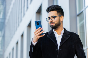 Handsome Indian businessman using smartphone typing text answering messages chatting online looking...