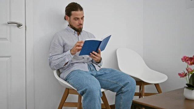 Young hispanic man reading book looking watch sitting on chair at waiting room
