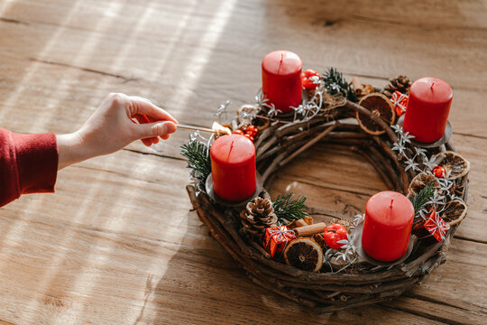 Advent wreath made with branches with four candles lit every sunday before christmas. Human hand holding a matchstick.