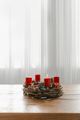 Advent wreath with four red candles on wooden dining table. White curtains background, cozy home to celebrate Christmas time.