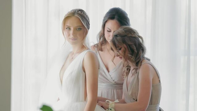 The bride and two girlfriends of the bride help to tie the ribbons of the dress. Preparation for the wedding, the morning of the bride. A beautiful bride with her bridesmaids