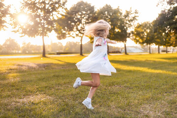 Happy girl child running with outstretched arms having fun in a city park, playing on a sunny day at sunset