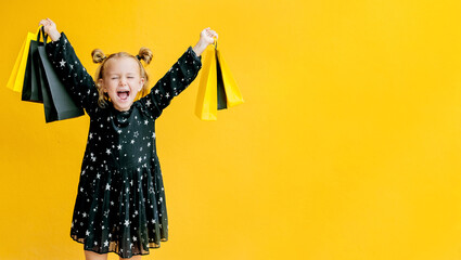 Banner smile child girl hold black package bags purchases isolated yellow background studio. Shopping buy online discount sale promotion. Cyber monday. Black friday. Toddler excited face kids emotions - obrazy, fototapety, plakaty