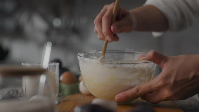 Woman is preparing dough for waffles, homemade pancakes, or pie at home. Wooden spoon in hand. Cook slowly stirs batter in glass bowl. Ingredients on table include wheat flour, eggs, sugar, and salt