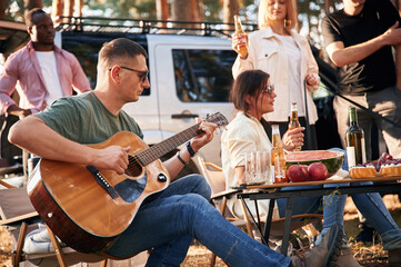 Man is playing guitar, by table. Group of friends are together in the forest
