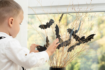 Halloween concept. Autumn holiday. The boy stands with his back near the window against the backdrop of a black vase with batting mice cut out of paper on dry branches. Close-up.