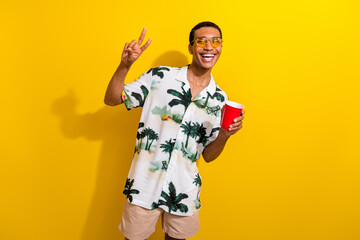 Photo portrait of attractive young man hold paper cup disco show v-sign wear trendy palms print clothes isolated on yellow color background