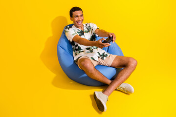 Full size photo of handsome young guy sit blue bean bag play video game wear trendy palms print outfit isolated on yellow color background