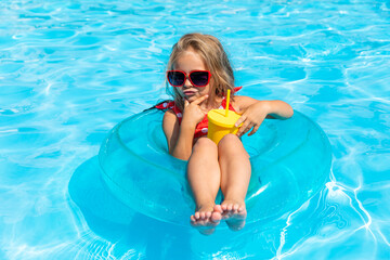 Pretty little girl swimming in outdoor pool and have a fun with inflatable circle and drinking...