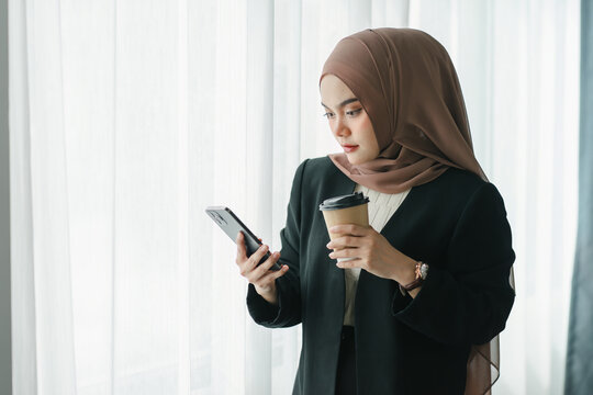 Asian Muslim businesswoman using a smartphone and enjoying coffee while standing in a modern office.