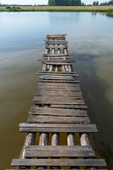 A wooden bridge made of logs and partially rotten boards. Being careful is dangerous. Water, lake, bush. Shore.