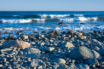 Behind the stone beach, the blue sea shines with its white waves.