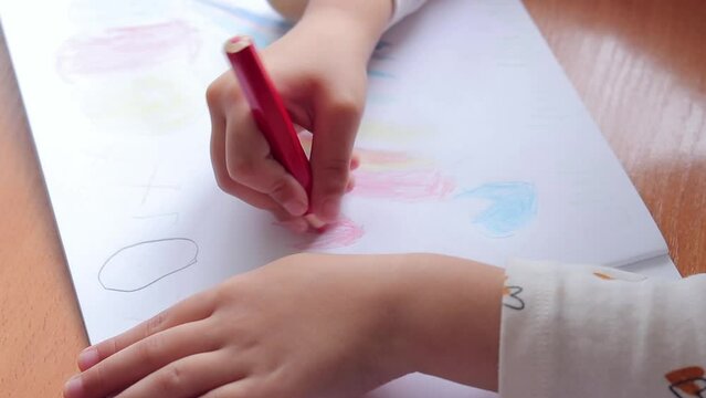 A preschool child draws with colored pencils in an album. Child's hand with a pencil close-up, selective focus. The girl draws hearts in red