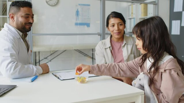 Waist up of Middle Eastern male child practitioner giving candies to Latin little girl and telling treatment recommendations to her mam while having checkup appointment in modern office