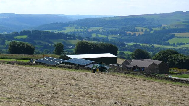 Farm tractor in the field on the hill slope above farm buildings raking hay with rotary rake into swaths or windrows. Peak District National Park, England, United Kingdom.