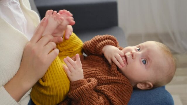 Playful young mother sitting on sofa together with pretty little infant son. Positive cute mom holding baby on lap while rest together on comfortable couch at home. Family, care, maternal love concept