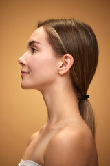 Profile view of tender, attractive young girl with ponytail, standing with bare shoulders isolated over light brown background. Concept of natural beauty, skincare, cosmetics and cosmetology
