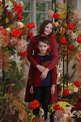 Mother with child with red rose flowers. Mom hugs her son, hugs with love. Coxy interior with flowers, family indoor. Fashion boy, cute kid and beautiful woman in red dress. Autumn decor, summer decor