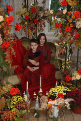 Mother with child with red rose flowers. Mom hugs her son, hugs with love. Coxy interior with flowers, family indoor. Fashion boy, cute kid and beautiful woman in red dress. Autumn decor, summer decor