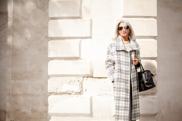 A  beauttiful woman with grey hair is walking arounв the old town. A stylidh woman is wearing a gray coat, a skirt and a bag and sunglasses