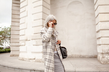 A  beauttiful woman with grey hair is walking arounв the old town. A stylidh woman is wearing a gray coat, a skirt and a bag and sunglasses