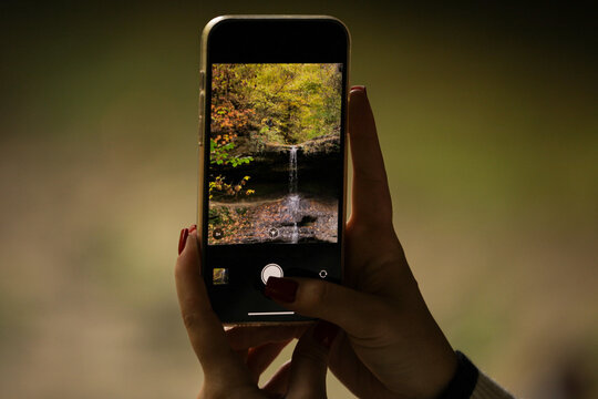 The picture of a young lady holding a phone and trying to take a picture of a beautiful autumn landscape, in the background is a waterfall and many colorful leaves, autumn colors.