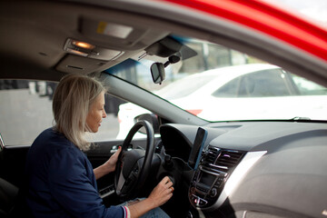 A  beauttiful woman with grey hair is driving car around the old town. A stylidh woman is wearing a jeans, blue jacket